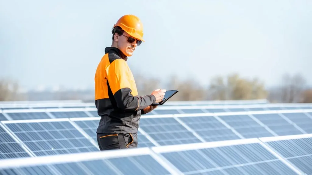 Solar installer checking panel output on a tablet beside a large panel array